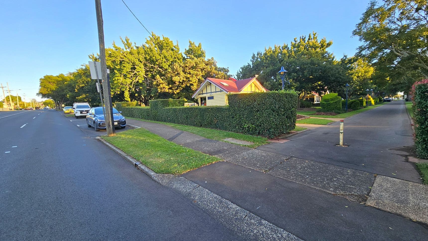 A suburban street with a footpath and a row of parked cars on the left. A blue car is closest, followed by a white van and a yellow car. A wooden house with a red roof is on the right, surrounded by a neatly trimmed hedge. The footpath leads to a paved area with a bollard. Trees with dense foliage line both sides of the street, casting shadows on the ground. A streetlamp is visible near the house. The road is wide with multiple lanes.