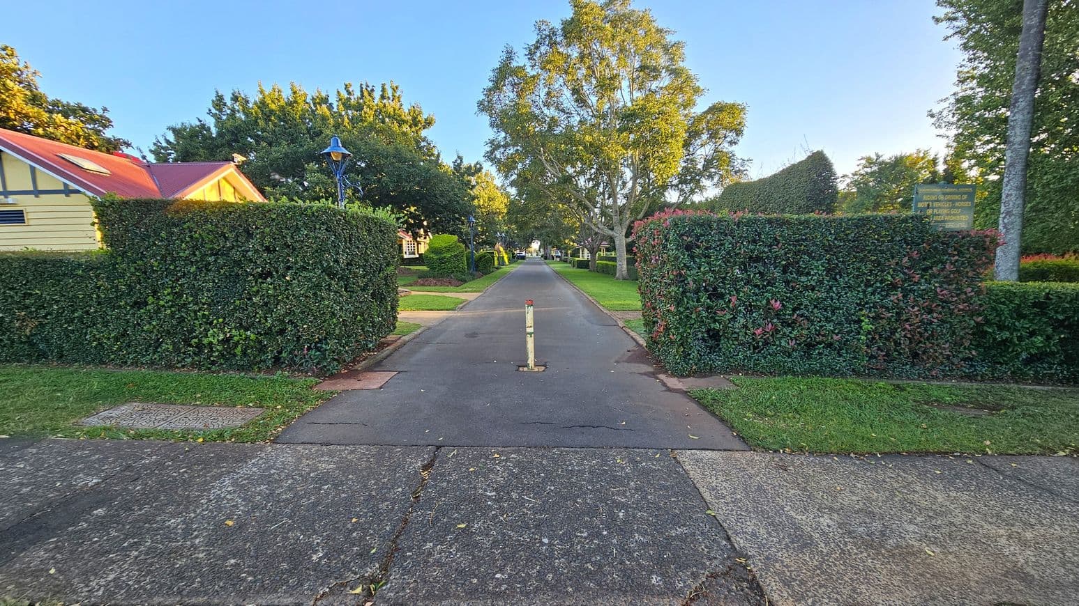 A paved path runs through the centre, flanked by neatly trimmed hedges. On the left, a building with a red roof and cream walls is partially visible. A blue lamp post stands nearby. On the right, a sign is mounted on a hedge, and a tall tree is visible. The path is bordered by grass, and a single bollard is positioned in the middle of the path. Trees line the background, creating a lush, green setting.