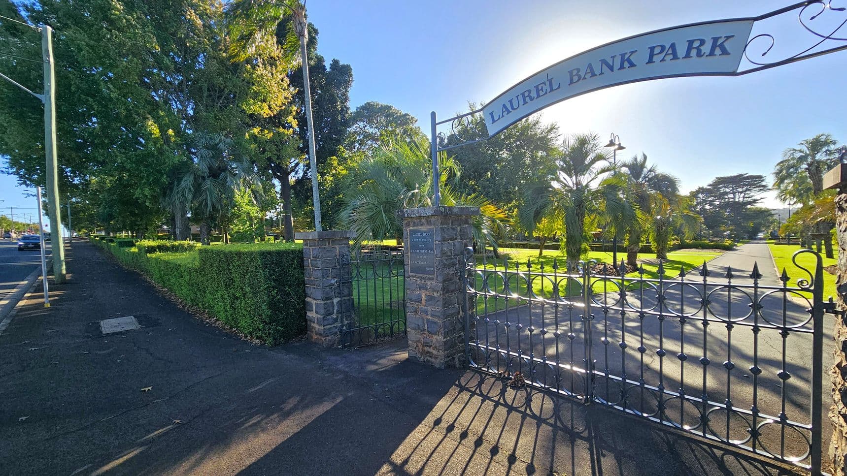A wrought iron gate with the sign "Laurel Bank Park" is open, leading into a park with neatly trimmed hedges and palm trees. The path inside the park is lined with grass and trees. On the left, a pavement runs alongside a road with a few cars. Tall trees provide shade along the pavement. The gate is supported by stone pillars, and sunlight casts shadows on the ground.