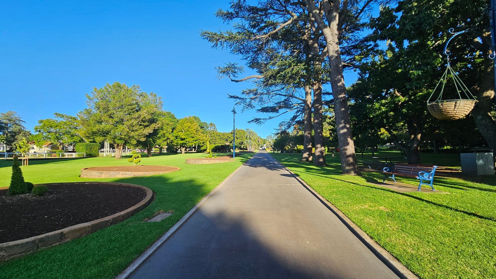 A paved path runs through a park, flanked by well-maintained grass. On the left, there are circular flower beds with soil, surrounded by lush trees. On the right, tall trees cast shadows over benches and a hanging basket. A lamppost lines the path, which stretches into the distance.