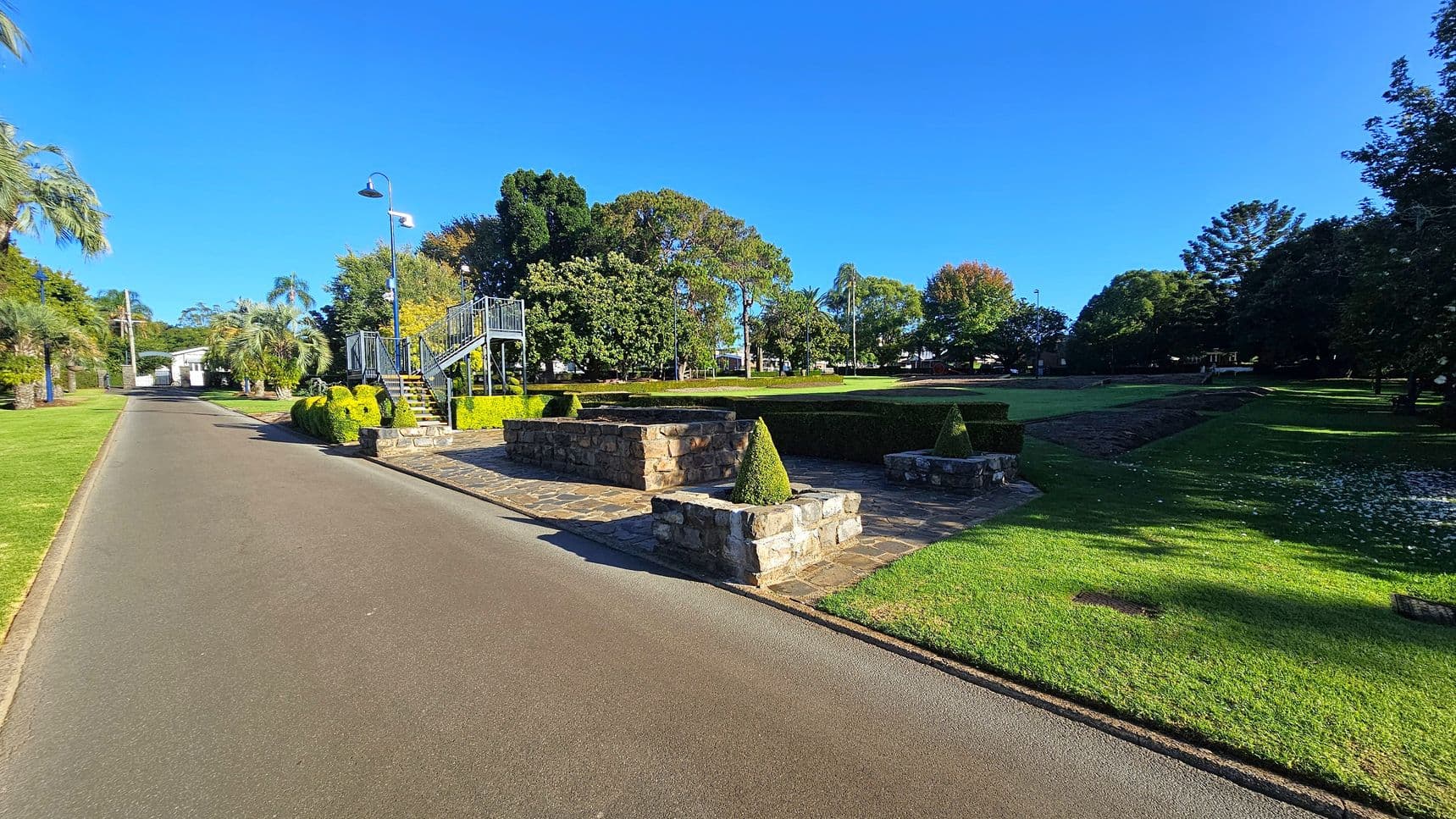 A paved path runs along the left side, bordered by neatly trimmed grass and palm trees. On the right, a landscaped area features stone planters with conical topiary bushes. A metal staircase with railings leads up to a small platform. In the background, a variety of trees provide a lush backdrop, with a mix of evergreen and deciduous foliage. The ground is covered with well-maintained grass, and a streetlamp is visible near the staircase.