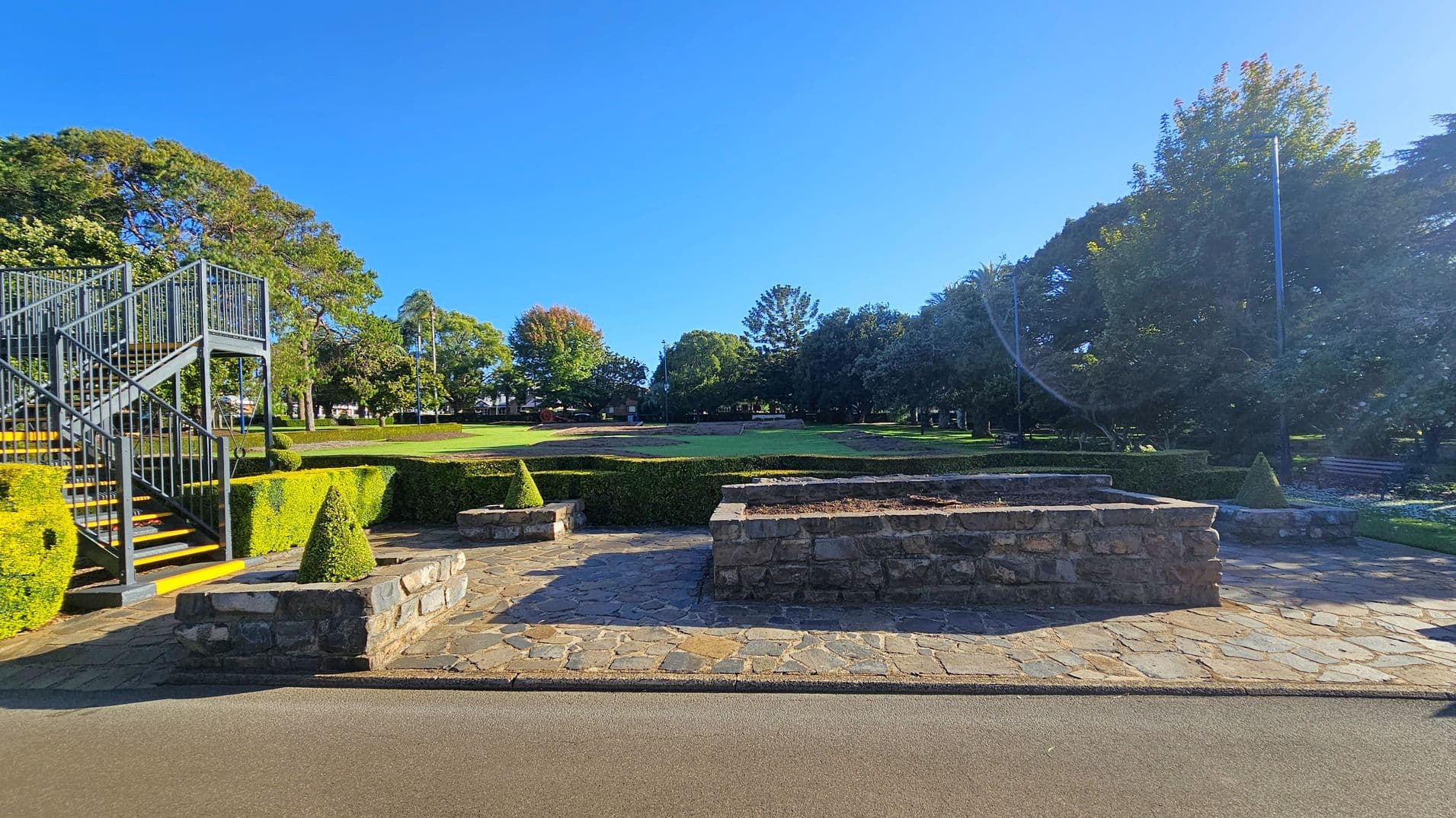 A park scene with a stone-paved area in the foreground, featuring low stone walls and neatly trimmed hedges. On the left, a metal staircase with yellow-edged steps leads upwards. The background is a grassy area bordered by various trees, including some with autumnal foliage. A pathway runs along the bottom edge of the image.