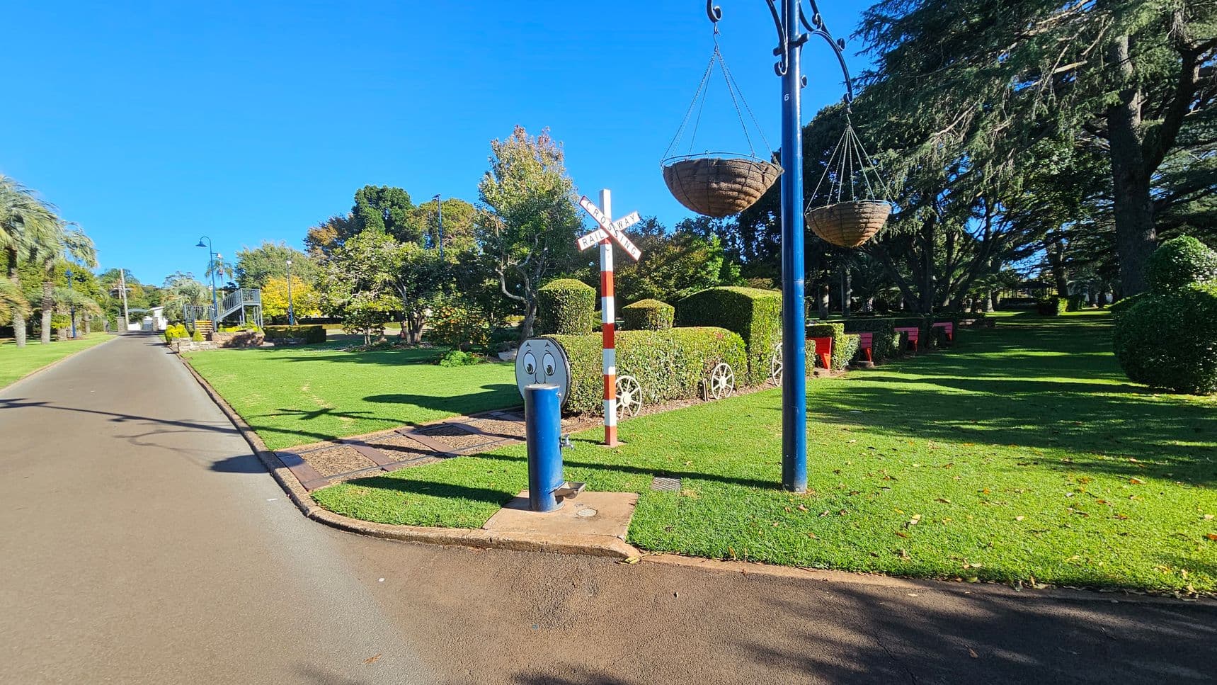 A paved path runs along the left side, bordered by well-maintained grass. On the right, there's a grassy area with a hedge shaped like a train, complete with wheels. A railway crossing sign stands nearby. Two hanging baskets are suspended from a blue pole. Trees and shrubs are scattered throughout the background, providing shade and greenery. A blue post with a cartoon face is near the path.