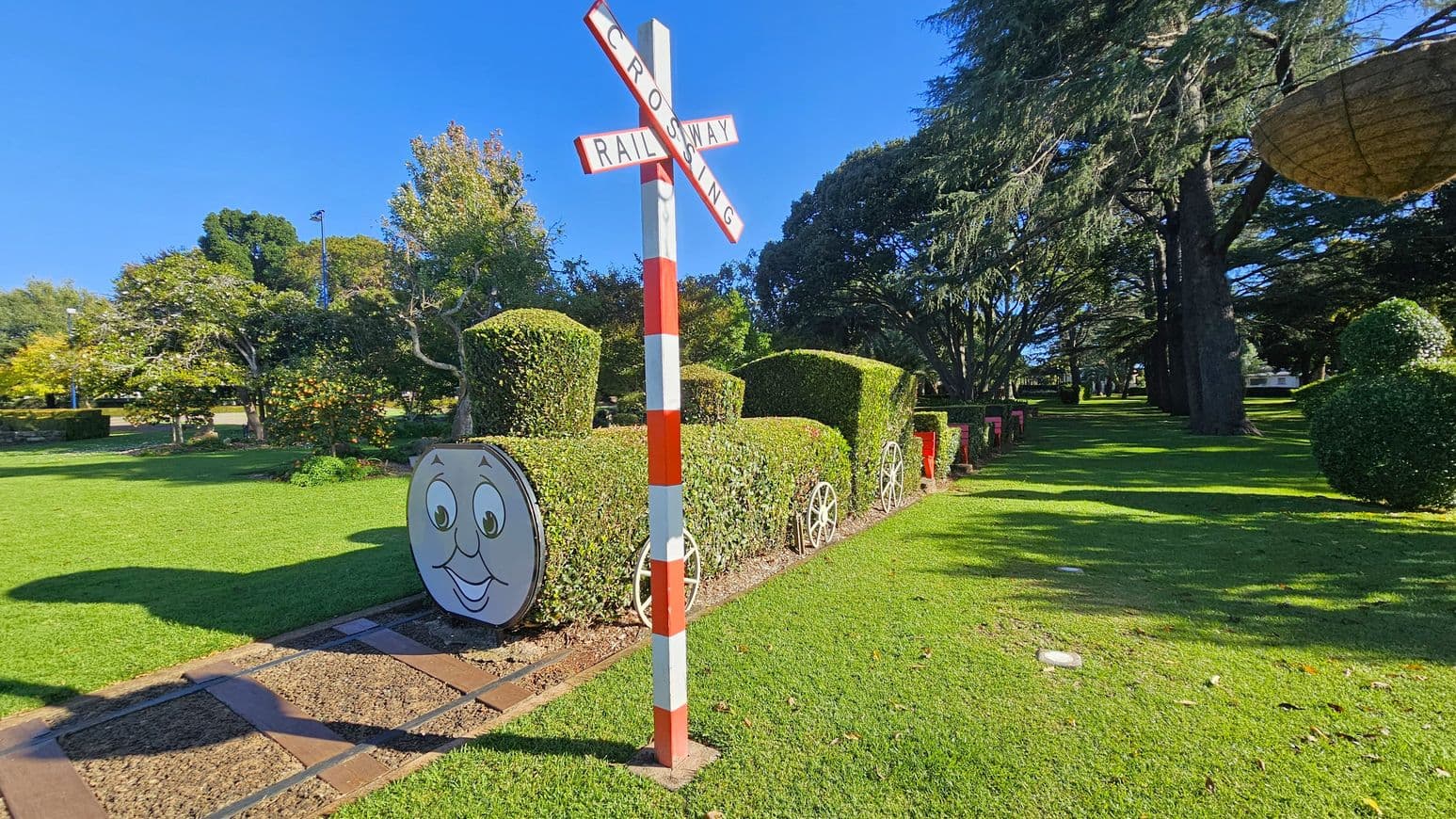 A garden scene with a railway crossing sign in the centre, featuring a red and white striped pole. To the right, a hedge shaped like a train with a smiling face on the front, complete with wheels and a chimney. The background has lush green grass and various trees, including a large tree casting shadows on the lawn. The sky is clear and blue.