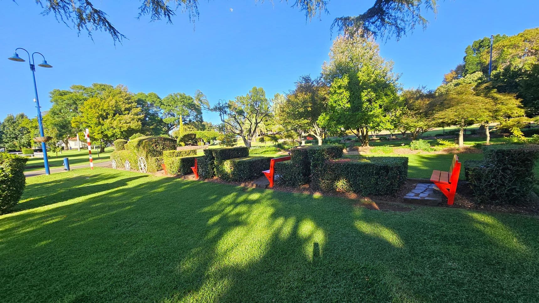 A well-maintained park with lush green grass and neatly trimmed hedges. In the centre, there are two bright red benches integrated into the hedges. To the left, a blue lamp post stands near a striped pole. The background features various trees with dense foliage, casting shadows on the ground. The sky is clear and blue, and the area is well-lit by sunlight.