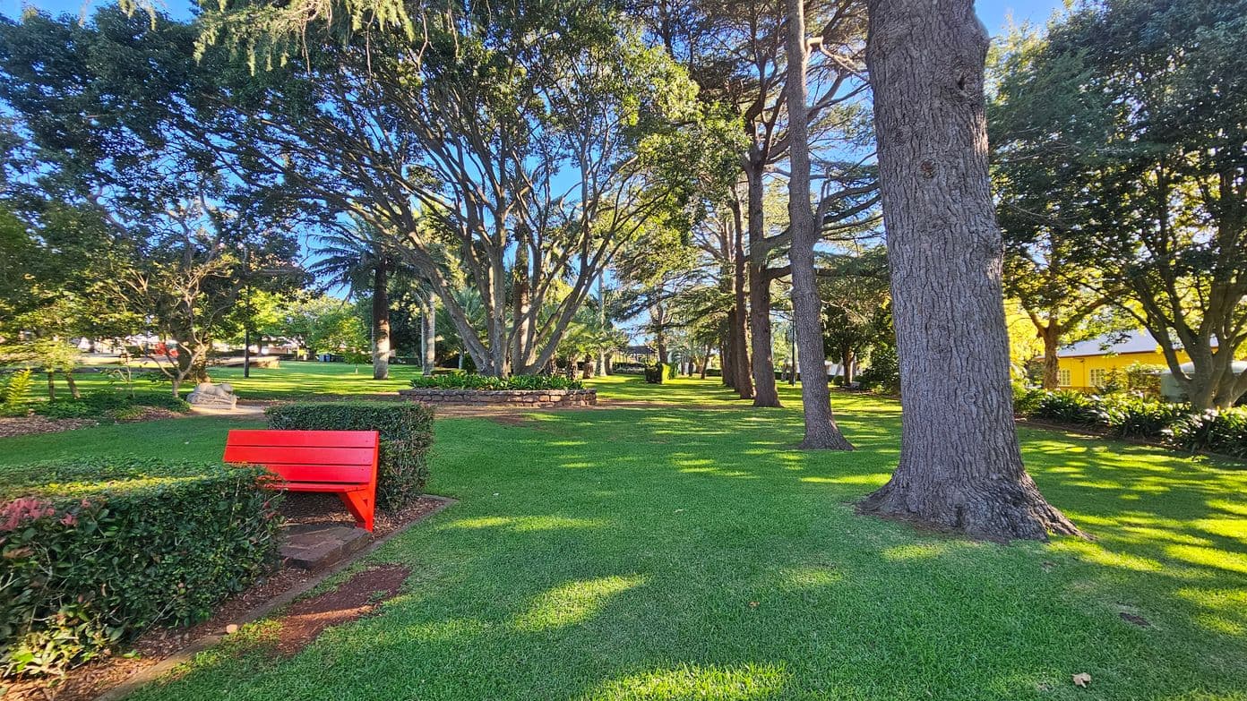 A park with lush green grass and tall trees scattered throughout. In the foreground on the left, there is a bright red bench next to a neatly trimmed hedge. The background features more trees and a yellow building partially visible on the right. Sunlight filters through the branches, casting dappled shadows on the ground.