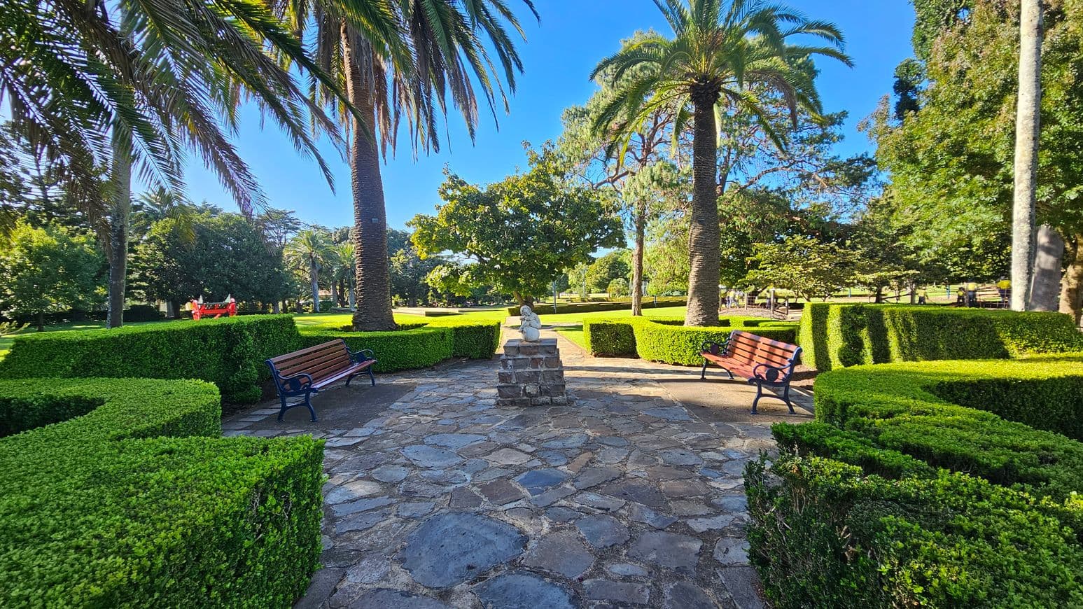 A stone pathway leads to a small statue on a pedestal in the centre, surrounded by neatly trimmed hedges. Two wooden benches with metal frames are positioned on either side. Tall palm trees rise on both sides, with lush green grass and various trees in the background. The scene is well-lit, suggesting a sunny day.