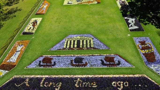A large grassy area features intricate floral displays. In the foreground, flowers spell out "A long time ago...". Above this, a triangular floral arrangement depicts three chariots. Further up, another triangular display shows a structure resembling a Greek temple. Surrounding these are rectangular floral panels with various historical and mythological scenes. The grass is neatly trimmed, and trees are visible on the right side.
