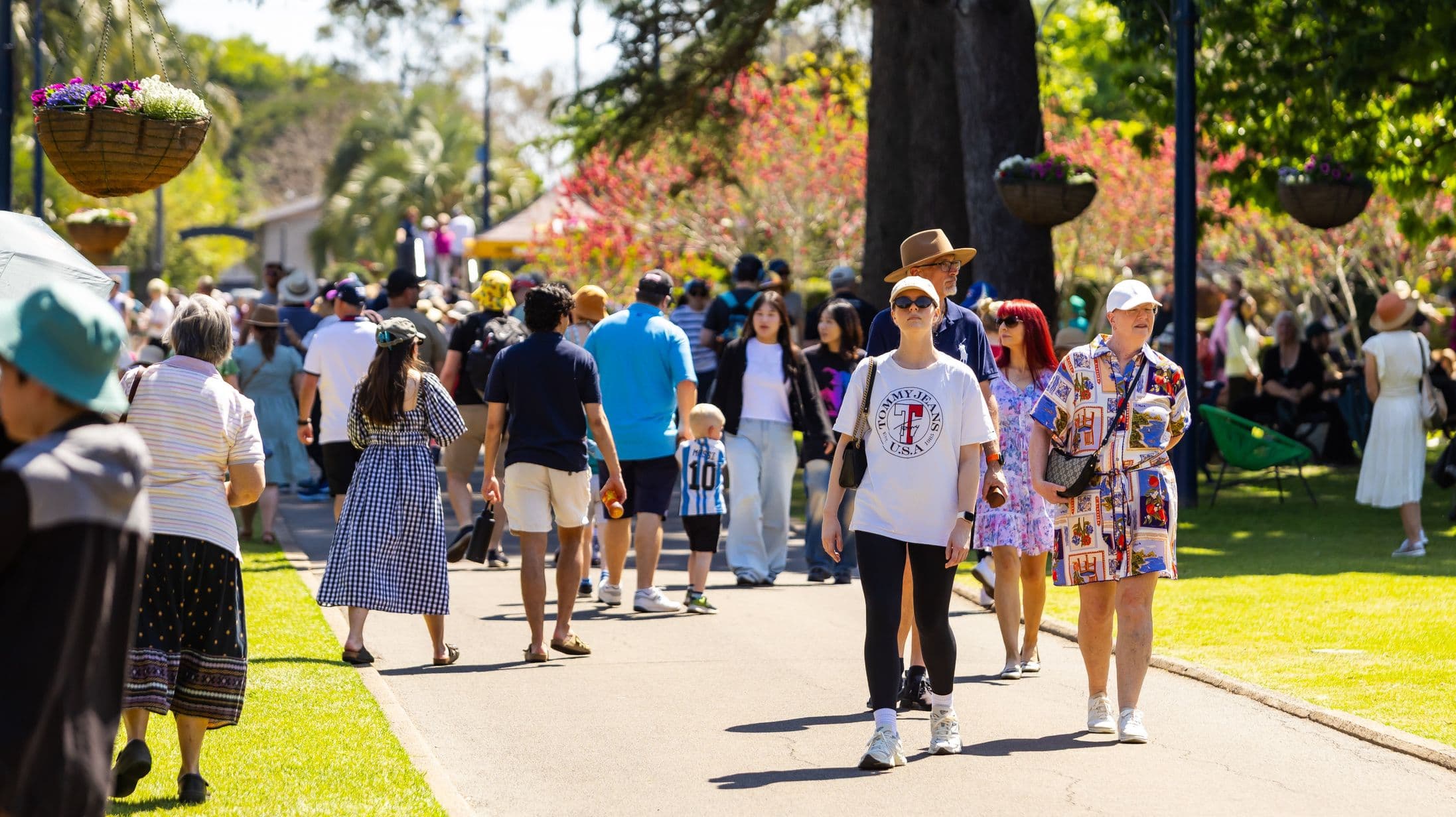 A bustling outdoor scene with people walking along a paved path. On the left, a woman in a blue and white checkered dress walks away, while others stroll in various casual outfits. On the right, a woman in a white cap and T-shirt walks alongside another in a colourful dress. Hanging baskets with flowers are visible above, and trees with pink blossoms line the path. The grass on either side is bright green, and people are seated in the background.
