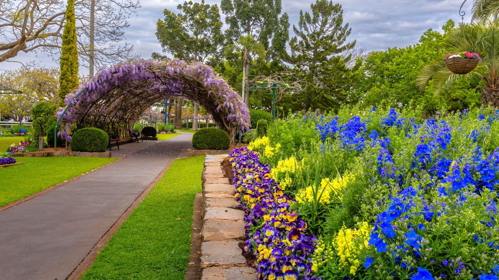 A garden path leads through a lush landscape. On the left, a wisteria-covered archway with benches underneath is surrounded by neatly trimmed bushes. On the right, vibrant flower beds with blue and yellow blooms line the path, bordered by a low stone wall. Tall trees and a hanging basket with flowers are in the background, adding to the garden's charm. The path is paved and flanked by well-maintained grass.
