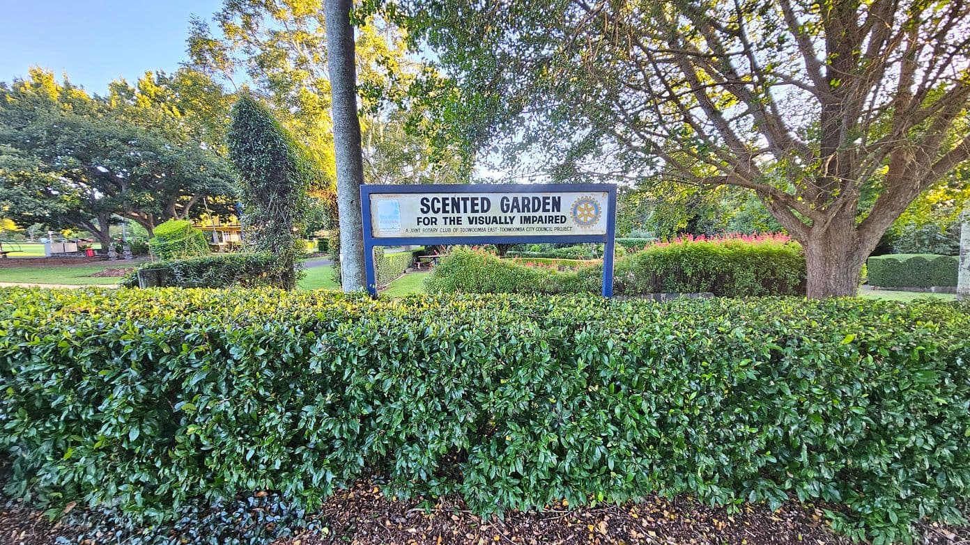 A sign reading "Scented Garden for the Visually Impaired" is centred in the image, supported by two posts. In the foreground, a neatly trimmed hedge runs across the bottom. The background features a variety of trees and shrubs, with sunlight filtering through the leaves. A path is visible to the left, leading deeper into the garden area.
