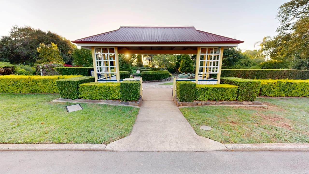 A small pavilion with a red roof and open sides stands in the centre, surrounded by neatly trimmed hedges. A concrete path leads up to the pavilion, flanked by green grass on either side. In the background, there are various trees and shrubs, creating a lush garden setting. The foreground features a paved road edge and a small plaque on the left side of the grass.
