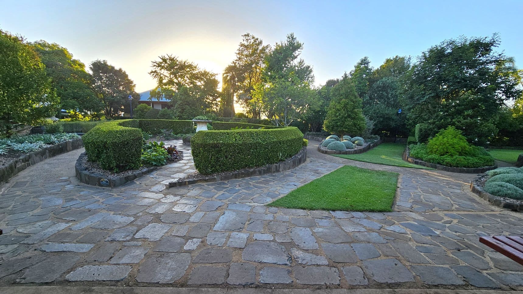 A well-maintained garden with a stone pathway winding through it. On the left, there are neatly trimmed hedges and a variety of plants. In the centre, a circular hedge encloses a small stone structure. To the right, there are more hedges and spherical shrubs. The background features tall trees with sunlight filtering through. The foreground shows a stone path with a patch of grass. A wooden bench is partially visible on the right.