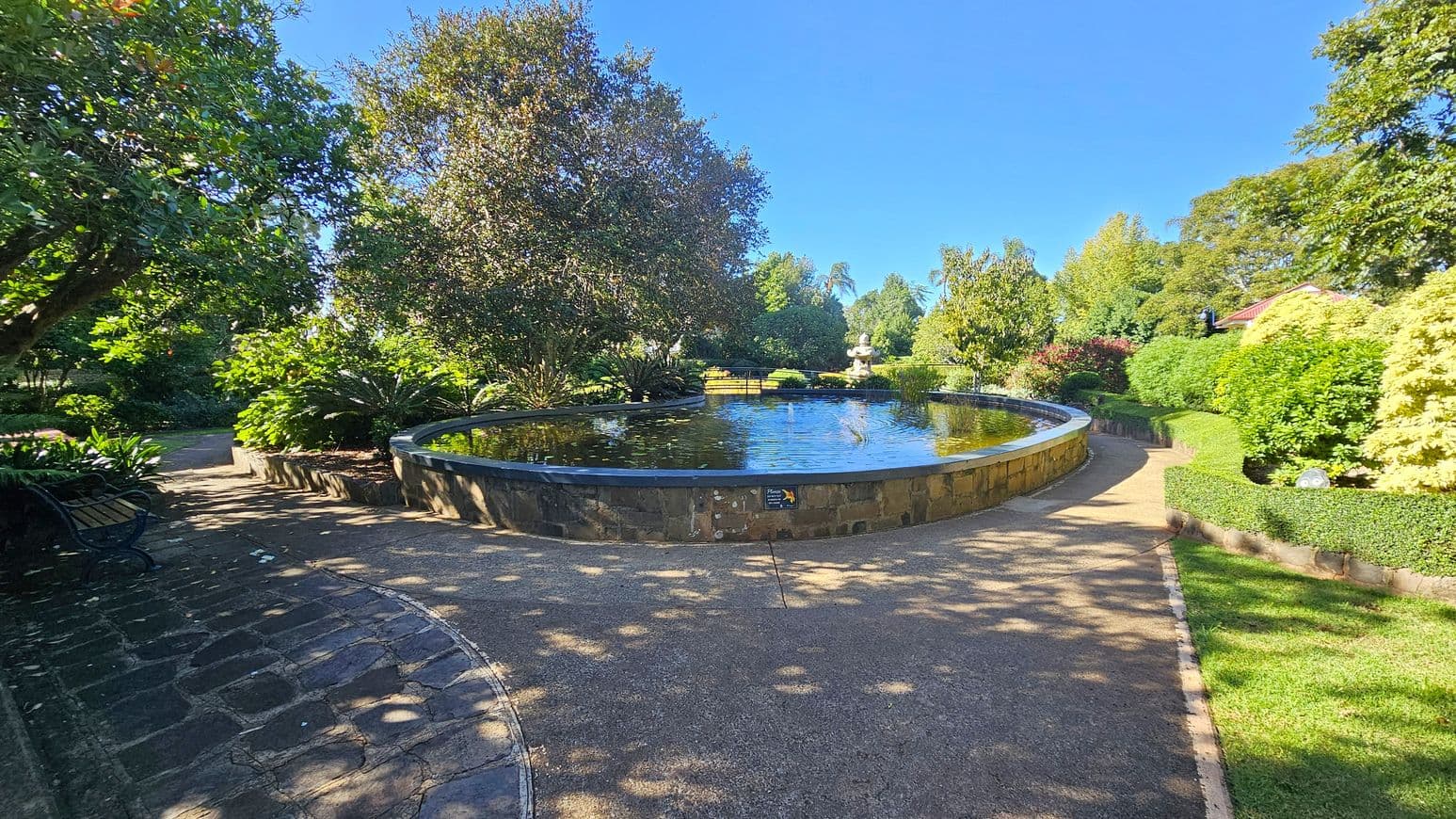 A circular pond is in the centre, surrounded by a stone wall. The water reflects the surrounding trees. On the left, a stone path curves around the pond, bordered by lush greenery and a bench. On the right, a gravel path is lined with trimmed hedges and more dense foliage. In the background, a statue is visible among the trees. The area is shaded by large trees, and the ground is a mix of stone and gravel.