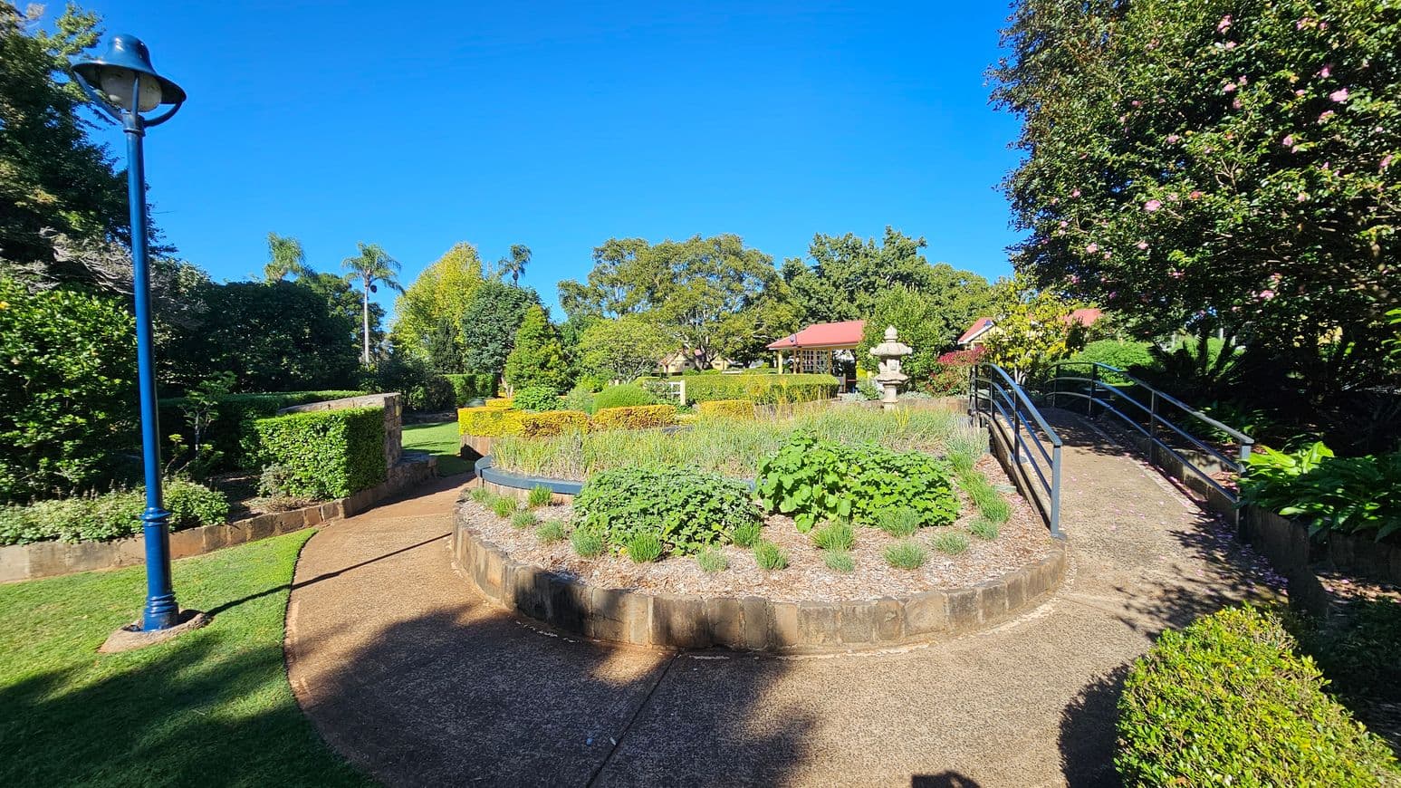A garden scene with a curved path in the foreground, bordered by lush greenery and shrubs. On the left, a blue lamp post stands beside a hedge. In the centre, a raised circular garden bed is filled with various plants. To the right, a metal-railed ramp leads upwards, flanked by flowering bushes. In the background, a red-roofed structure and a stone lantern are partially visible among the trees.