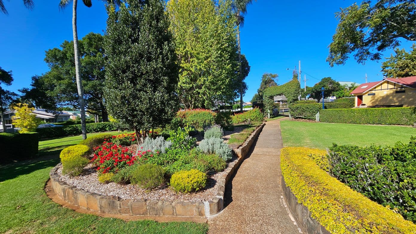 A well-maintained garden with a variety of plants and flowers, including red blooms and green shrubs, bordered by a stone edging. A narrow path runs through the garden, flanked by neatly trimmed hedges. On the right, a yellow building with a red roof is visible. Tall trees and a palm tree are in the background, with a clear blue sky above. The grass is lush and green, adding to the vibrant scene.