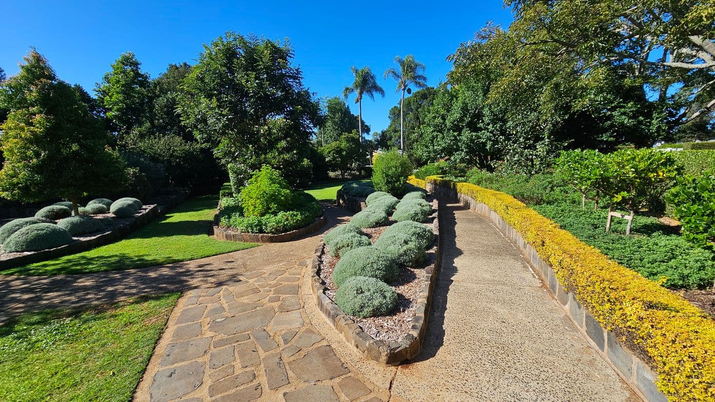 A well-maintained garden with a stone pathway in the centre. On the left, there are neatly trimmed round bushes and a variety of trees. On the right, a raised garden bed with small shrubs and a low yellow hedge borders the path. In the background, tall palm trees and dense greenery are visible. The ground is a mix of stone paving and grass.