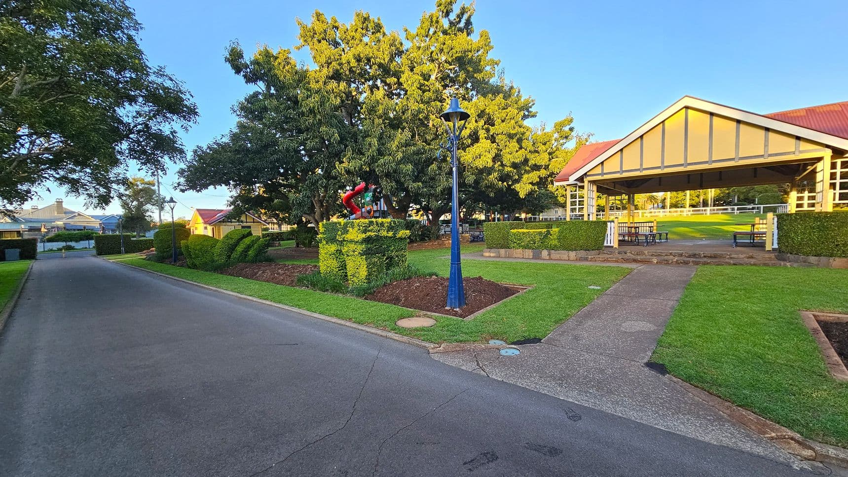 A paved road runs along the left side, bordered by lush green grass and trees. In the centre, a blue lamppost stands near a neatly trimmed hedge with a red sculpture on top. To the right, a pavilion with a red roof and open sides houses picnic tables. The area is surrounded by well-maintained lawns and hedges, with a large tree providing shade in the background.