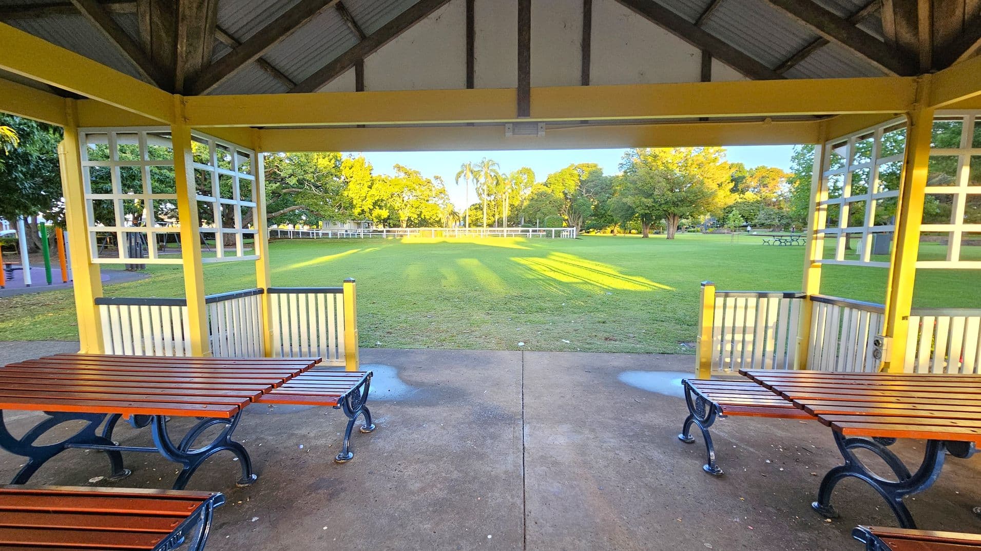 A park shelter with a wooden roof and yellow beams. Two wooden picnic tables with metal frames are on a concrete floor. The shelter opens to a large grassy field with trees in the background. Sunlight casts long shadows on the grass. A white fence runs along the edge of the field, and there are more trees and a few picnic tables in the distance.