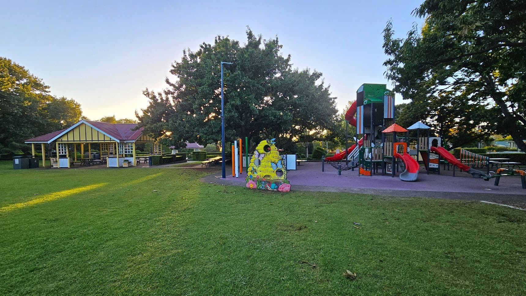 A playground with a grassy foreground. On the left, there's a pavilion with a red roof and yellow accents, surrounded by trees. In the centre, a colourful bee-themed decoration stands near a lamppost. On the right, a playground structure features red slides and climbing areas, set on a purple surface. Tall trees are in the background, providing shade.