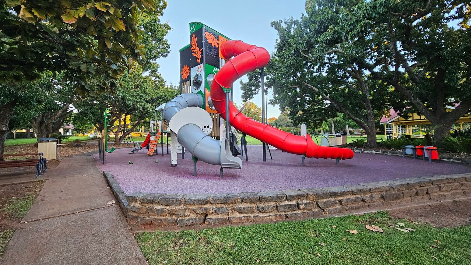 A playground with a large play structure featuring a red spiral slide and a grey slide. The structure has colourful panels with floral designs. Surrounding the playground are large trees providing shade. The ground is covered with purple safety flooring, and there's a stone border separating it from the grass. Benches and bins are visible to the left, and a building with a red roof is in the background.