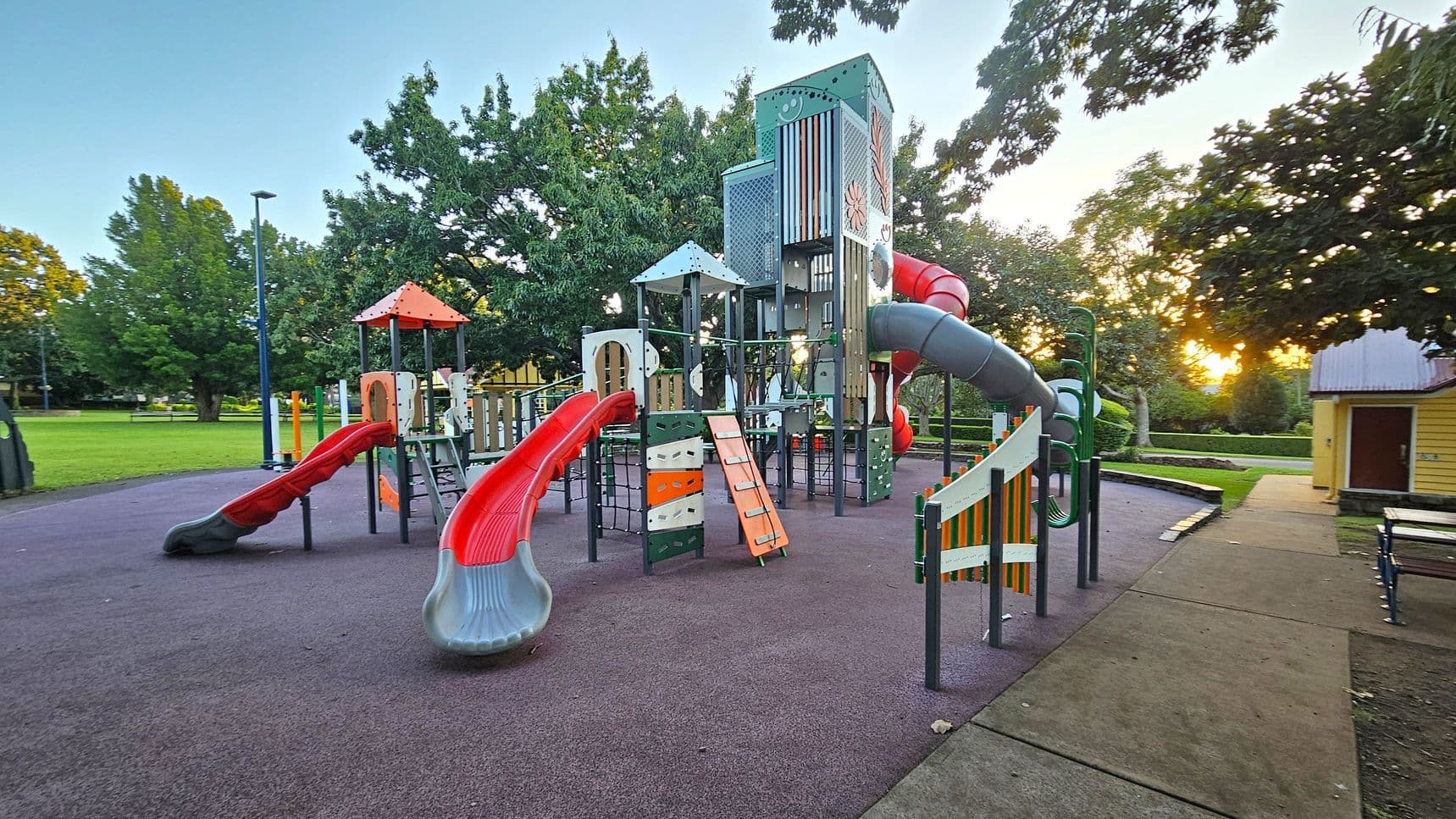 A playground with various equipment including slides, climbing structures, and tunnels. The ground is covered with a purple safety surface. On the left, there are red slides and a small climbing wall. In the centre, a tall structure with multiple levels and a tube slide. On the right, a path leads to a yellow building. Trees surround the area, and the sun is setting in the background, casting a warm glow.
