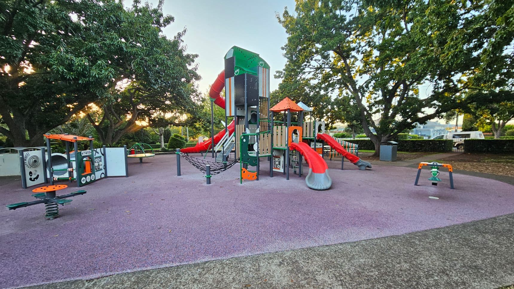 A playground with various equipment on a purple rubber surface. On the left, there's a small play structure resembling a store with a steering wheel and a spring-mounted seat. In the centre, a large climbing structure with multiple red slides and a rope bridge. On the right, a small green and orange seesaw. The background features large trees with dense foliage, and a fence with a bin nearby. The ground is a mix of purple rubber and concrete.