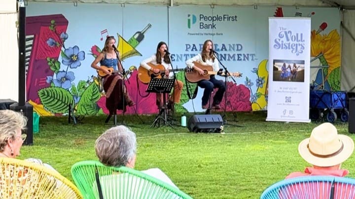 Three musicians are seated on a stage, each playing a guitar. Behind them is a colourful mural with flowers and a sign that reads "People First Bank" and "Parkland Entertainment." A banner on the right displays "The Volt Sisters." In the foreground, several people are seated on vibrant, woven chairs on a grassy lawn, watching the performance. The setting appears to be outdoors under a tent.