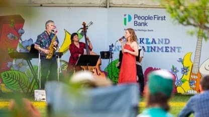 A band is performing on an outdoor stage. On the left, a man plays a saxophone, while a woman next to him plays a double bass. In the centre, a woman in a red dress sings into a microphone. The backdrop features the logo of "People First Bank" and the words "Parkland Entertainment" with colourful illustrations of musical instruments. The foreground shows blurred heads of audience members, suggesting they are seated on the grass.