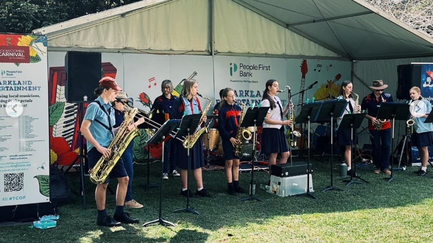 A group of musicians is performing under a large tent. On the left, a sign with colourful graphics and text stands beside a speaker. The musicians, mostly wearing uniforms, are playing various instruments including saxophones and clarinets. Music stands are in front of them. The background features a banner with text and logos. The ground is covered with grass, and the tent's ceiling is visible above.