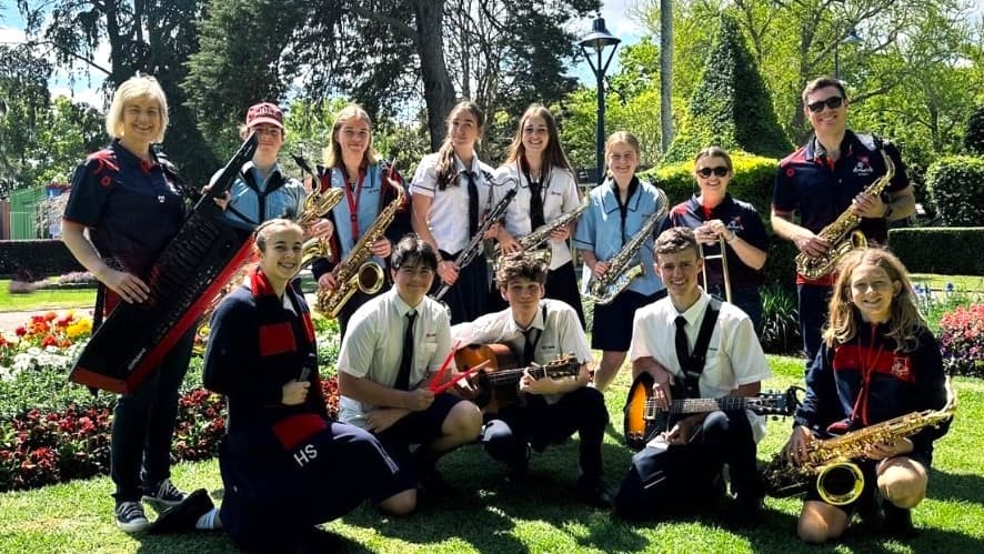 A group of people, mostly in school uniforms, are gathered on a grassy area. Some are standing, holding musical instruments like saxophones and a keyboard, while others are kneeling with guitars. They are surrounded by flower beds and trees, with a lamppost visible in the background. The scene is well-lit, suggesting a sunny day.