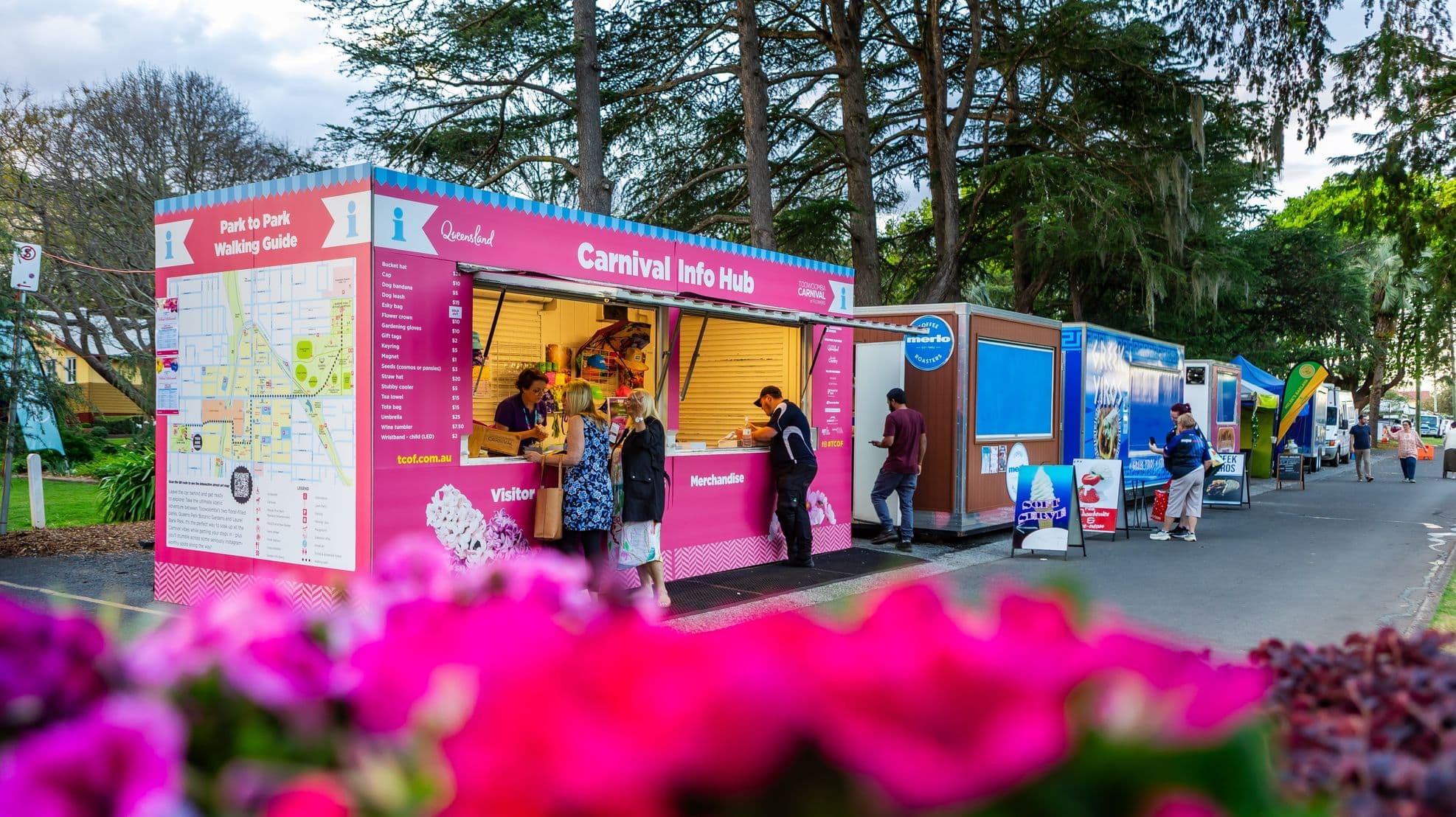 A vibrant carnival scene with a pink "Carnival Info Hub" booth on the left, featuring a map and merchandise list. People are gathered around, engaging with staff. To the right, a row of food stalls and trailers, each with distinct signage and colours. In the foreground, bright pink flowers are slightly out of focus. Tall trees form the background, creating a lively and festive atmosphere.