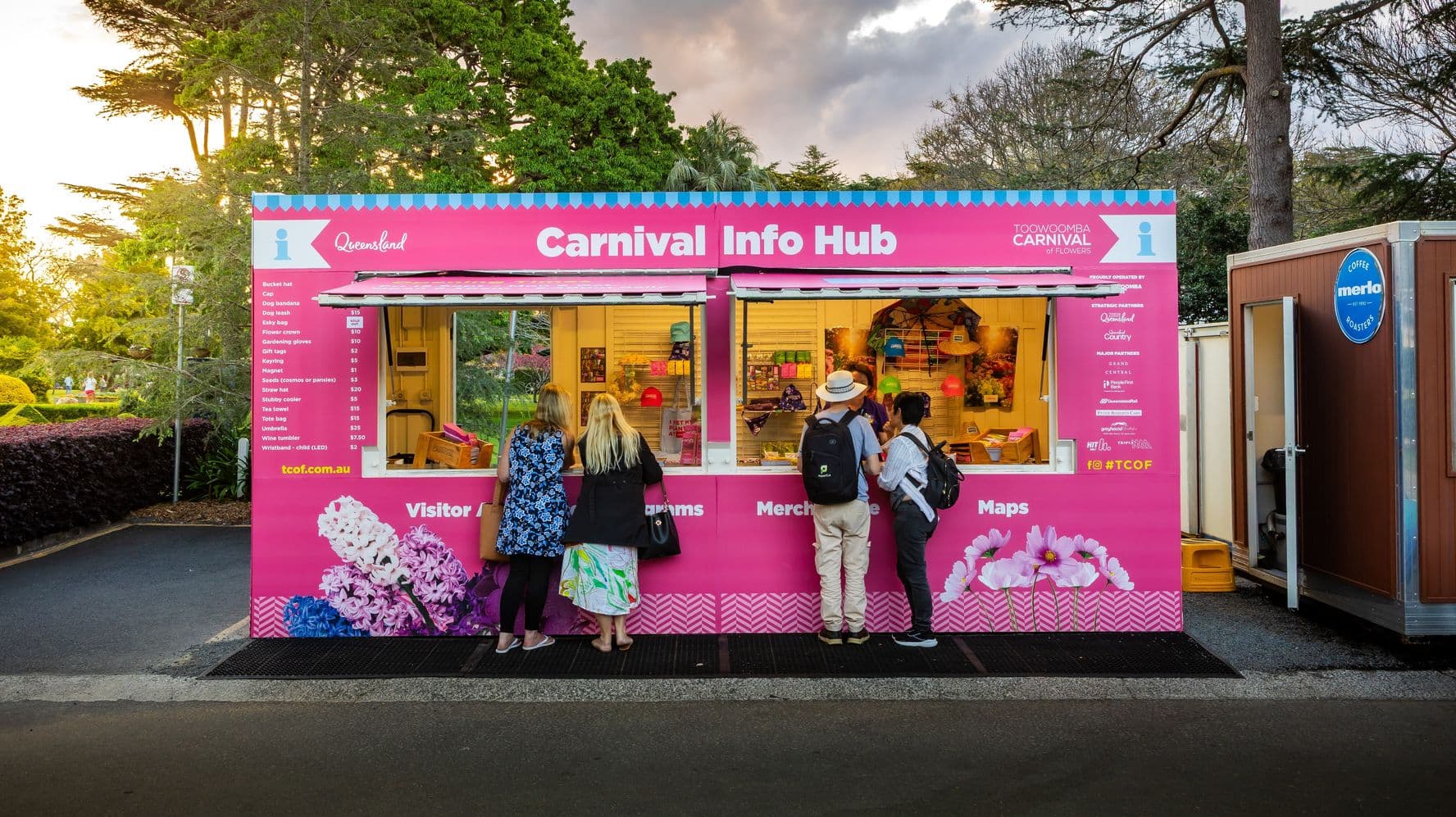 A bright pink Carnival Info Hub with two service windows is in the centre. Four people stand in front, looking at merchandise and information. The hub is decorated with floral images and text about the Toowoomba Carnival of Flowers. To the right, a brown coffee booth with an open door and a blue sign. Trees and greenery are in the background, with a paved path in the foreground.