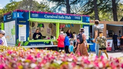 A vibrant outdoor scene with a Carnival Info Hub booth in the centre. The booth is blue and green, staffed by people assisting visitors. Several individuals, including children, are gathered around, engaging with the staff. In the foreground, colourful flowers are in bloom, adding a lively touch. Trees are visible in the background, providing a natural setting. The ground is paved, and the atmosphere appears busy and festive.