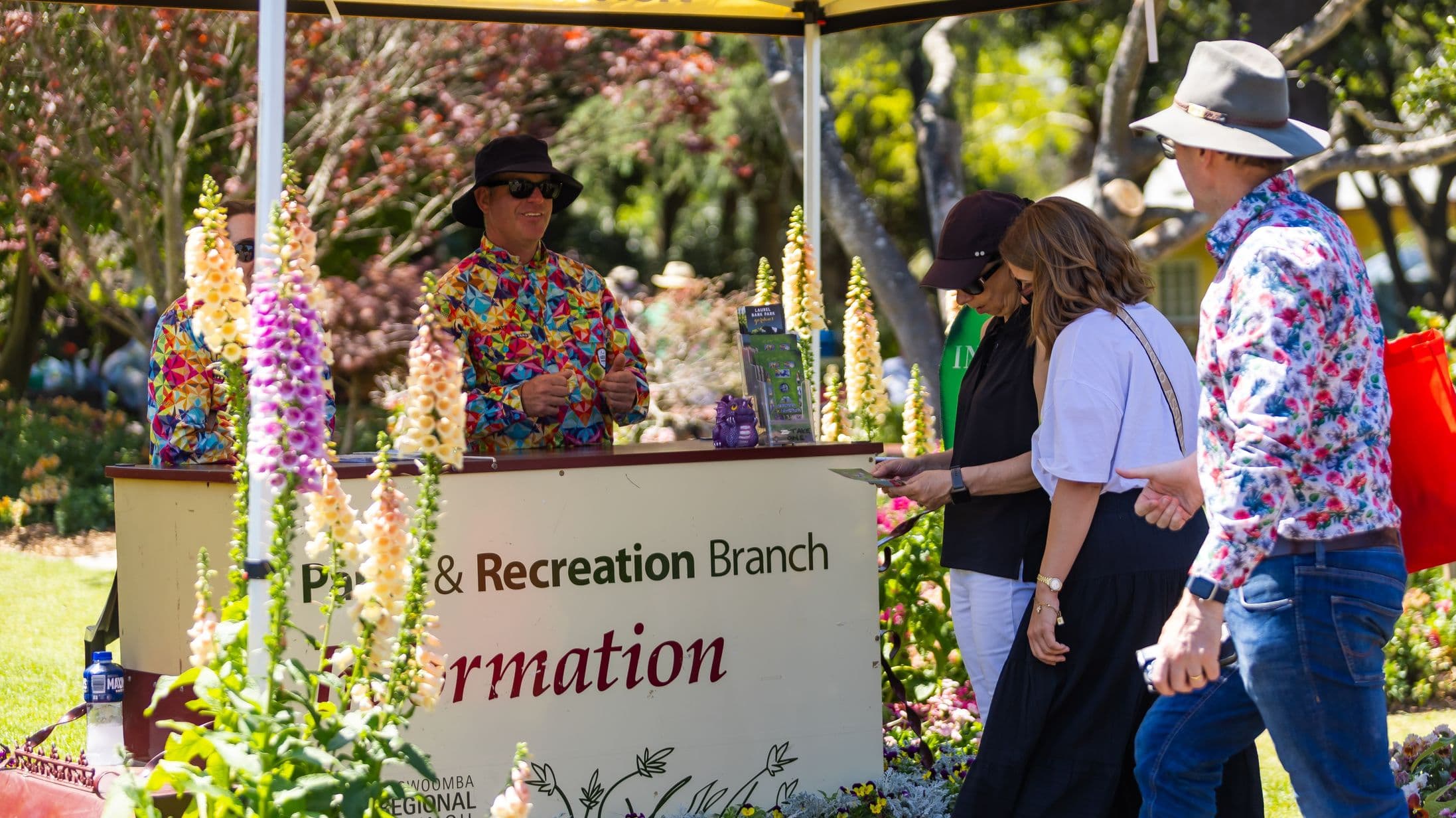 A man in a colourful, geometric-patterned shirt and black hat stands behind an information booth with a sign reading "Parks & Recreation Branch Information". In front of the booth, several people are gathered, including a person in a floral shirt and grey hat. Tall, multicoloured flowers are in the foreground. The background features trees and greenery, suggesting a park setting.