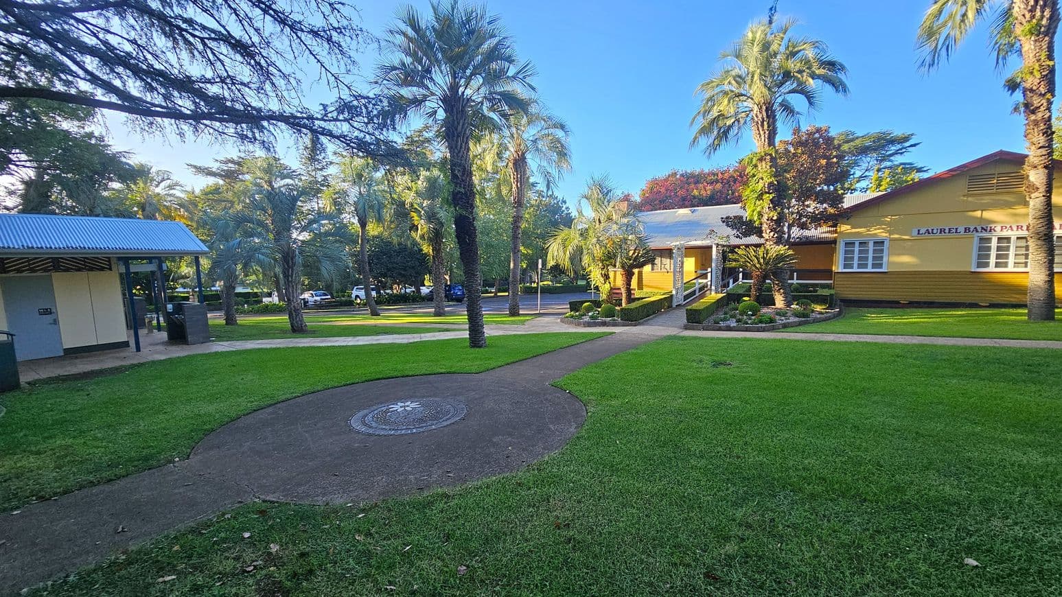 A park scene with a well-maintained lawn and a path leading through the centre. On the left, there's a small building with a corrugated metal roof. In the middle, tall palm trees are scattered around. On the right, a yellow building with a sign reading "Laurel Bank Park" is visible, surrounded by neatly trimmed hedges and small plants. Cars are parked in the background, partially obscured by trees.