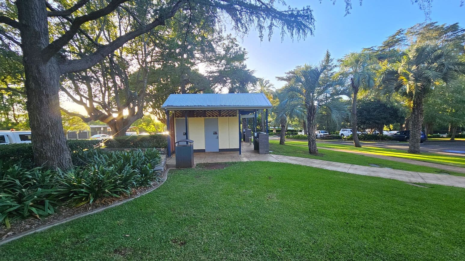 A small public restroom building with a corrugated metal roof is in the centre, surrounded by a well-maintained grassy area. On the left, large trees with thick trunks and dense foliage provide shade. On the right, there are several palm trees lining a paved path. In the background, a few parked cars are visible along a road. The sunlight filters through the trees, casting shadows on the grass.