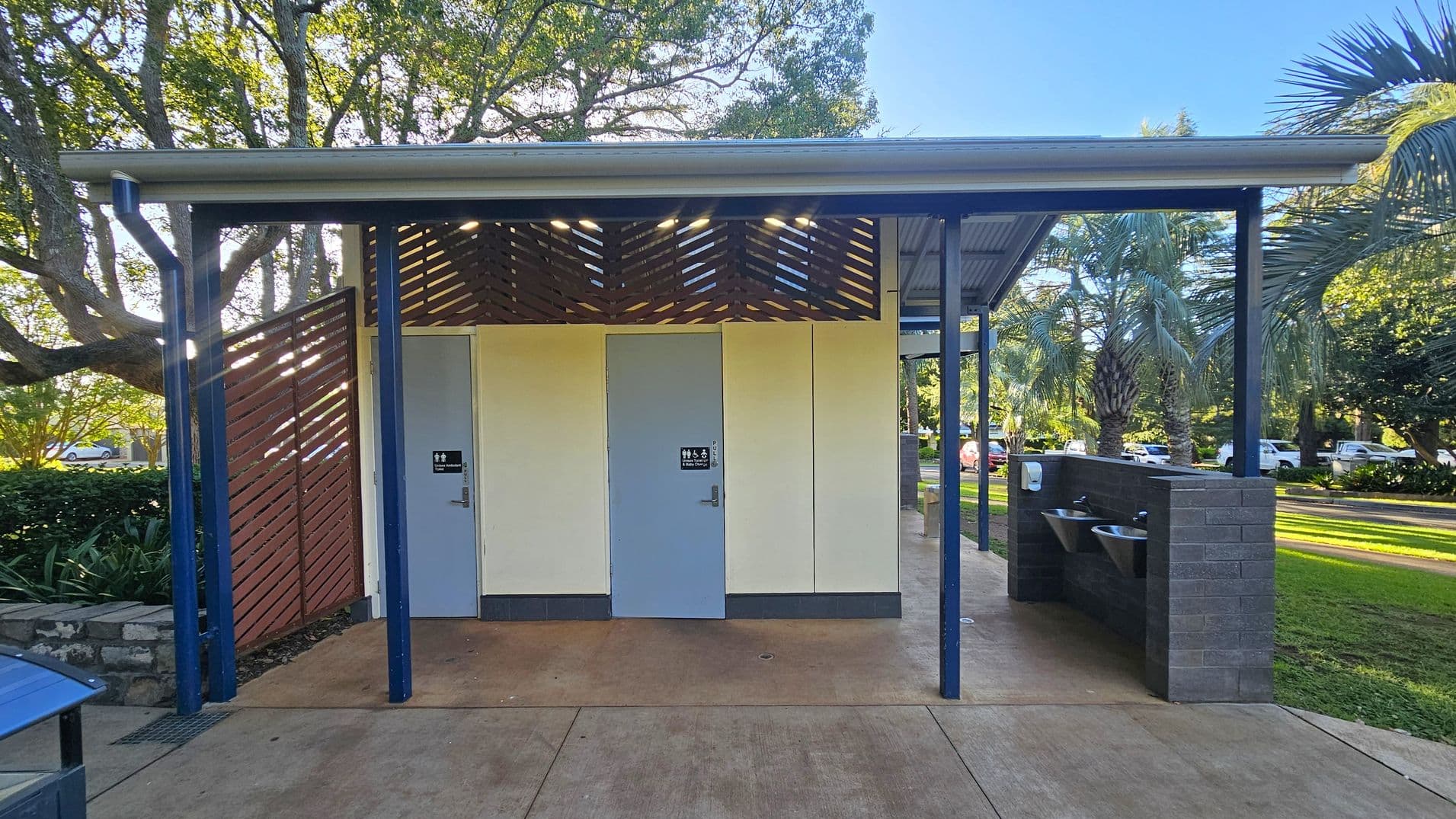 A public restroom facility with two doors in the centre, each marked with signage. The structure has a slatted wooden design above the doors and is supported by blue metal beams. On the right, there are outdoor sinks with a soap dispenser mounted on a brick wall. The ground is paved, and the area is surrounded by lush greenery, including large trees and palm plants. Cars are visible in the background, parked along a road.