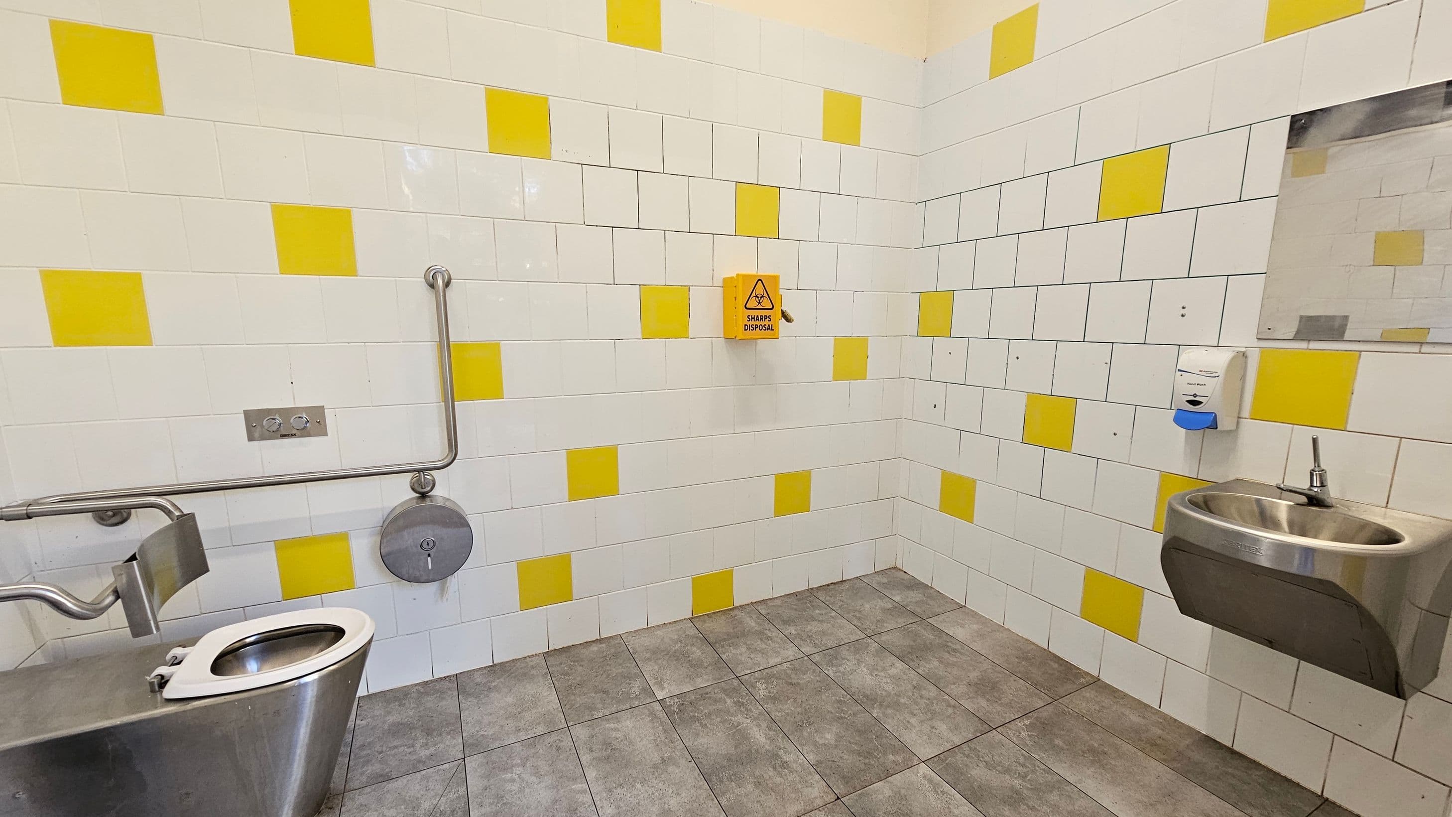 A public restroom with a stainless steel toilet on the left, equipped with a grab rail and a toilet paper holder. The walls are tiled with a pattern of white and yellow tiles. In the centre, a yellow sharps disposal box is mounted on the wall. On the right, a stainless steel sink with a soap dispenser above it is visible, along with a mirror. The floor is covered with grey tiles.