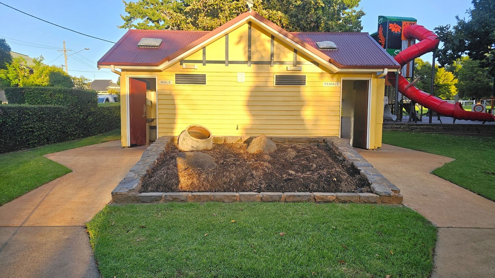 A small building with a red roof and yellow walls, featuring two entrances labelled "Male" on the left and "Female" on the right. In front, there's a garden bed with rocks and a large pot. The ground is paved with concrete paths leading to each entrance, surrounded by green grass. On the right, a red spiral slide is part of a playground structure. Trees and hedges are visible in the background.