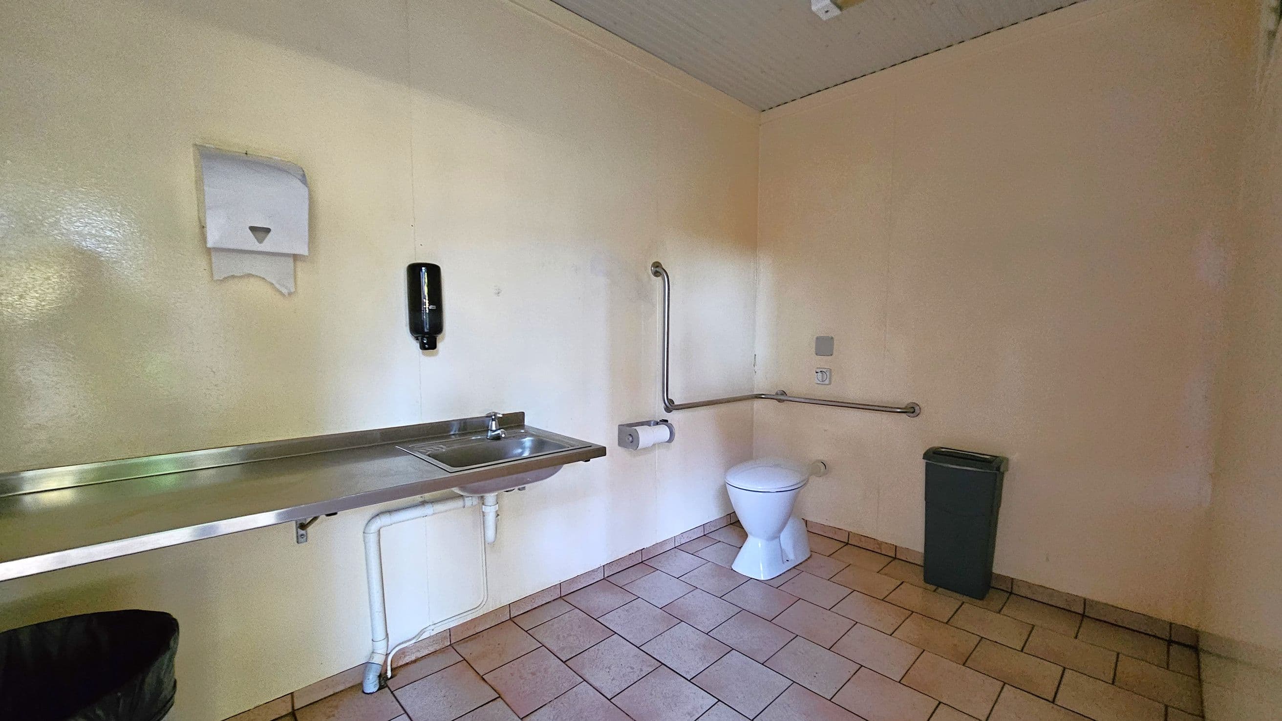 A bathroom with beige walls and a tiled floor. On the left, a stainless steel sink with a paper towel dispenser above and a soap dispenser mounted on the wall. Below the sink, visible plumbing pipes. On the right, a white toilet with a grab bar beside it and a toilet paper holder. A grey waste bin is positioned near the toilet. The ceiling is light-coloured with a small fixture.