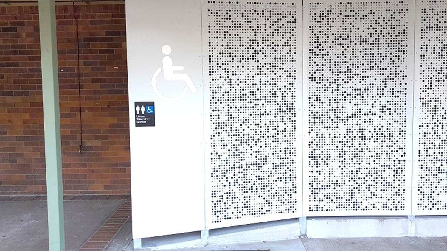 A wall with a perforated metal panel on the right, featuring a pattern of small, evenly spaced holes. To the left, a sign indicates accessible toilets, with symbols for male, female, and wheelchair access. The background has a brick wall partially visible. The floor is concrete, and a vertical green beam is on the far left.