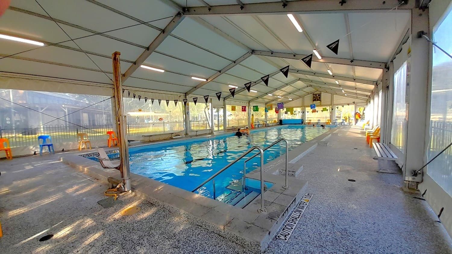 The indoor pool is visible from the inside and has a large white permanent marquee with transparent retractable awning walls, strings of banners with vibrant pennants strung between the ceiling and the blue water surface, a silver access ramp on both sides, colorful monobloc chairs, long benches, and numerous reminders written on the stone flooring and pool sides about the water level.
