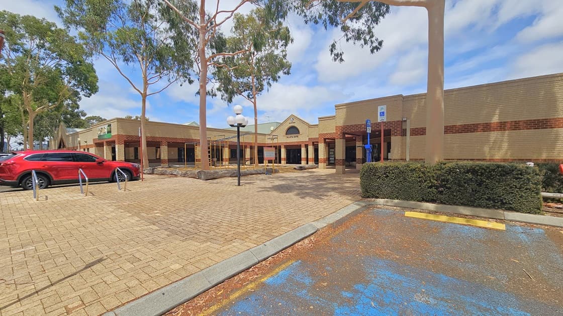 A low-set structure made of brown and cream bricks, with the building concourse featuring the same brown brick pattern. The facility banner hangs on the left side. Humps on the road, marked by yellow and black lines, are on the left. A red car is parked nearby. An accessible parking area, equipped with a step-free ramp with handrails is situated on the right. Lamp posts and trees are visible in front of the building.
