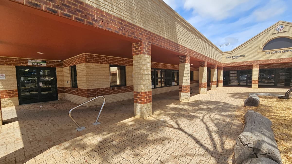 A low-set structure made of brown and cream bricks, featuring a pitched roof displaying the city and the facility's logo. To the left, a glass door marked "Entry" includes a stainless handrail. On the right, a large wooden structure borders the park.