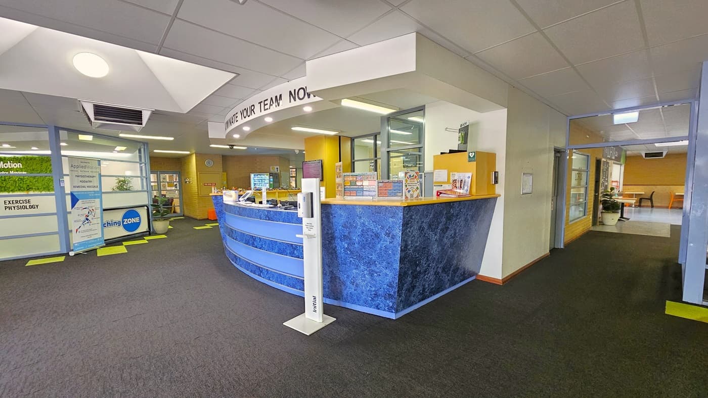 The reception with a high countertop, featuring a contrasting blue design. Bold letters on top read "Nominate Your Team Now.". Posters, banners, a computer, and a staff member are visible. An alcohol dispenser is positioned in front. To the left is the Exercise Physiology counter bearing their banner. To the right is a lobby leading to the cafe. The ceiling is white, and the grey carpet is accented by neon green tiles.