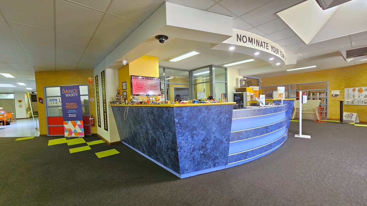 The reception, hosting a high countertop with a contrasting blue design. Bold letters on top read "Nominate Your Team Now." Posters, banners, a computer, and a staff member are visible. An alcohol dispenser is positioned in front. To the left, a large banner is displayed. Lobbies leading to the cafe are visible on both sides of the reception. The ceiling is white, and the grey carpet is accentuated by neon green tiles.