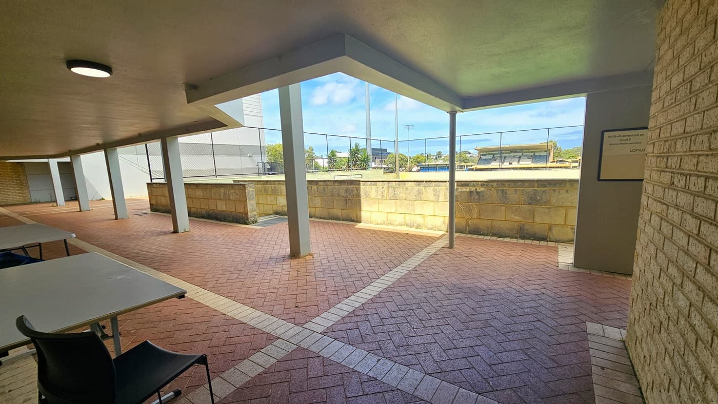 To the left is a wooden ramp leaning against the wall. To the right is a door made of heavy metal. Poster reminders are affixed to the wall and a green fire exit sign is placed on top. The ceiling has a cream rough structure. On the right is a concrete barrier made of bricks.