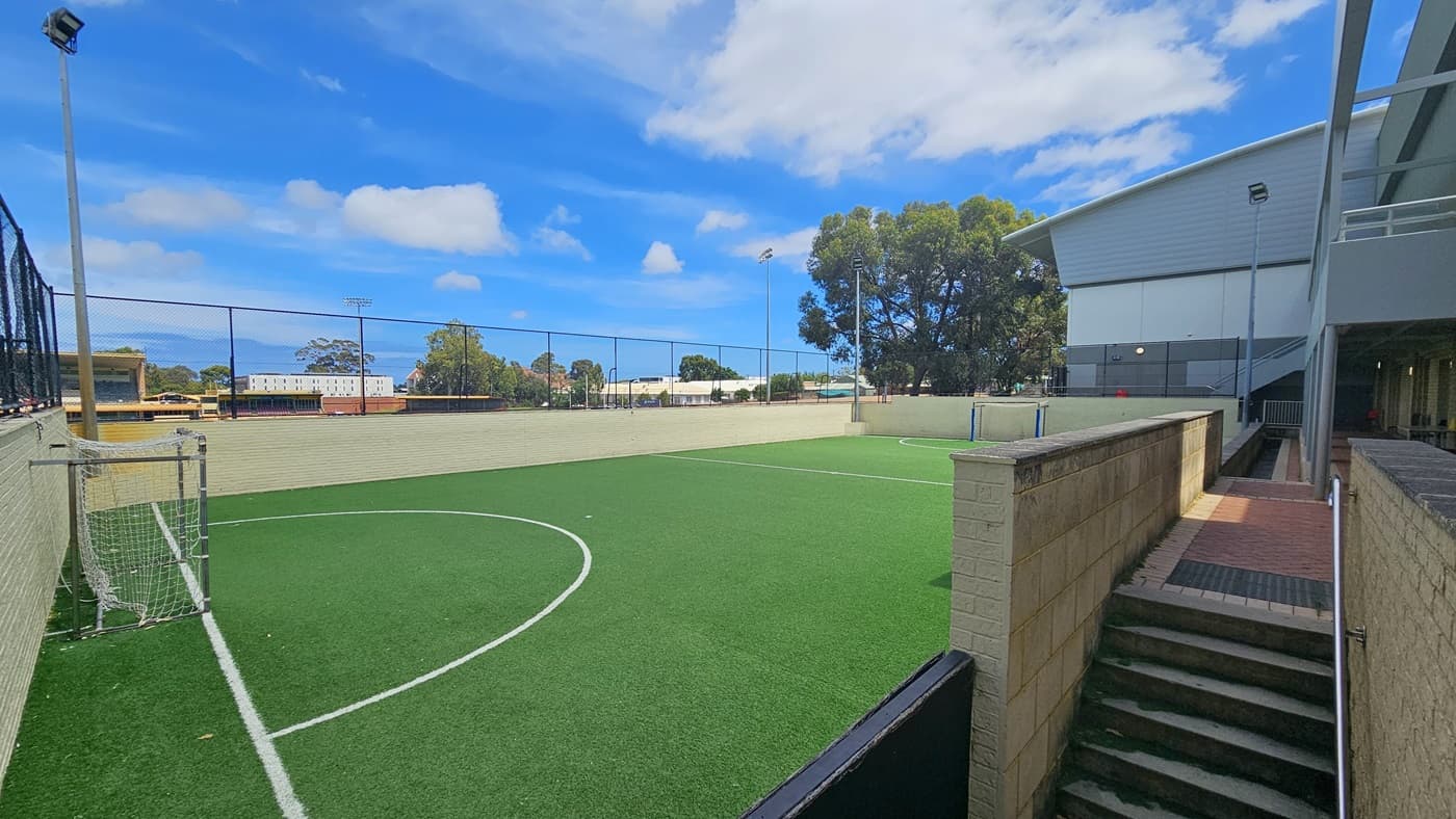 To the right are concrete stairs equipped with handrails. The rebound court, made of synthetic turf, is a spacious area with markings for the game. On both sides are goals equipped with nets. The entire space is surrounded by a chain fence. A tall floodlight is installed on the left. Further, commercial buildings are visible.