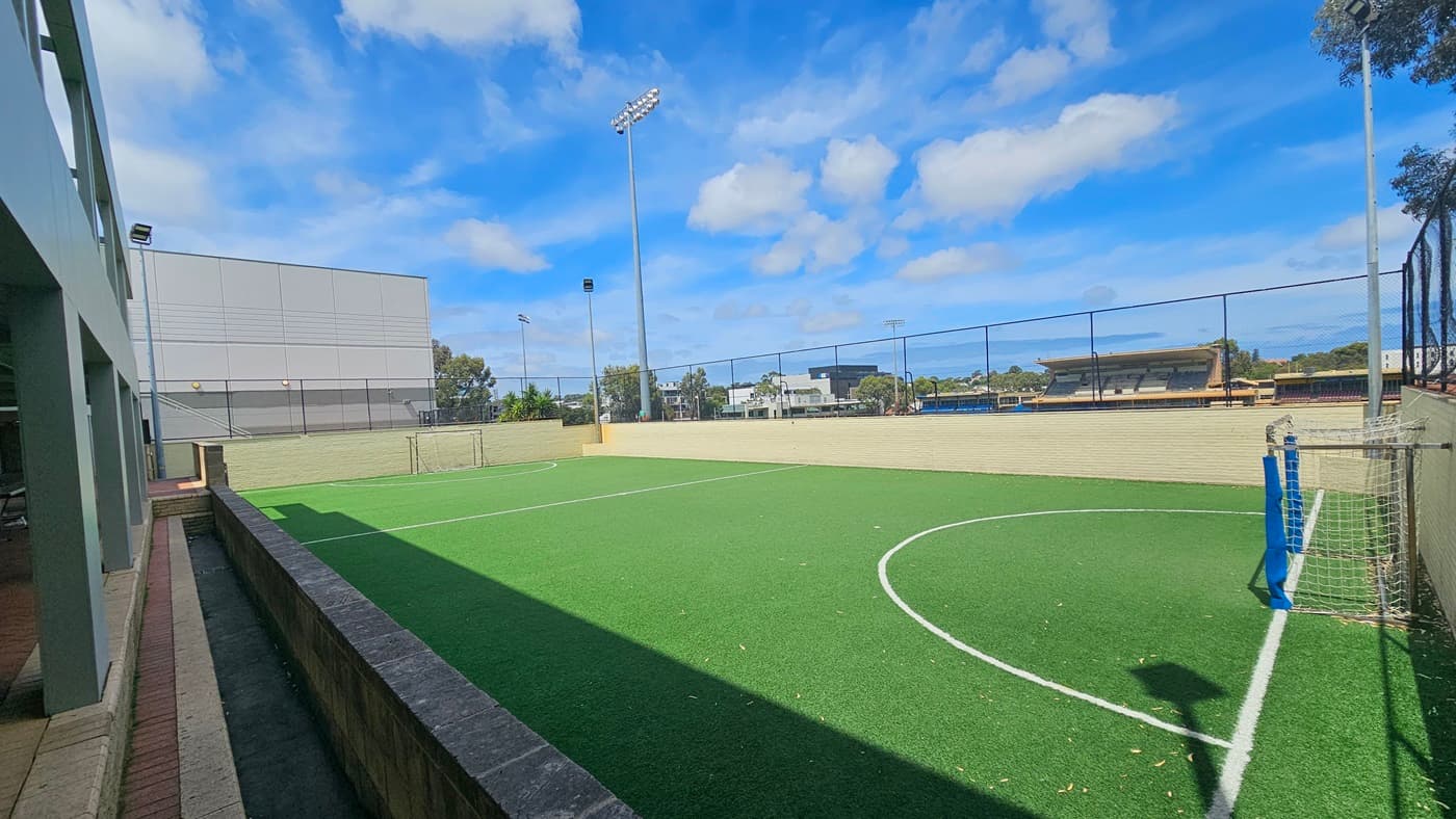 The rebound court, made of synthetic turf, is a spacious area with markings for the game. On both sides are goals equipped with nets. The entire space is surrounded by a chain fence. Tall floodlights are installed on the left. Further, commercial buildings are visible. On the left is a low wall that extends to the other side of the court.